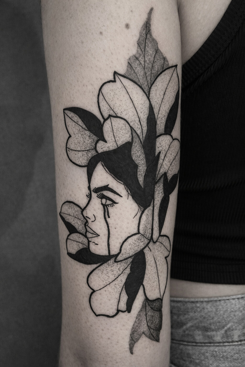 Black ink illustrative tattoo of a woman’s face combined with flowers, fine line and dotwork tattoo style, minimalist feminine tattoo